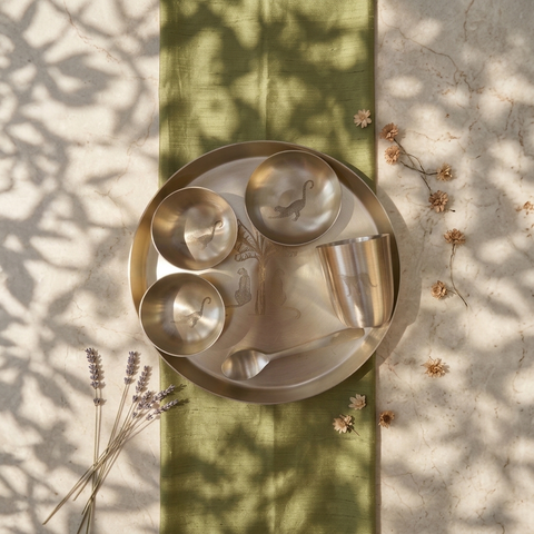 A set of matte metallic Indian tea and coffee cups, a matching spoon, and a large serving platter arranged on a green linen runner. The setup is styled with dried lavender sprigs and small flowers on a sun-dappled marble surface.