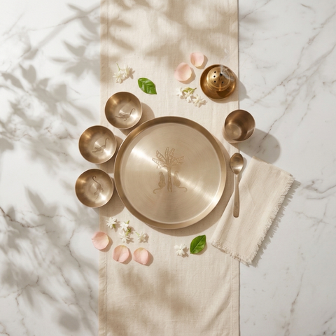 A set of brass tableware, including a large round platter, four bowls, a spoon, and a small decorative vessel, arranged on a cream linen runner over a white marble surface with soft shadows and flower petals.