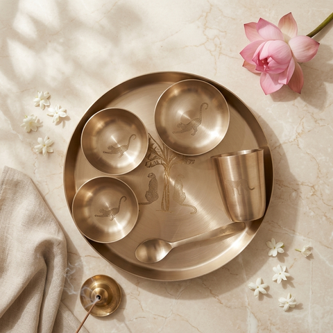 A Kansa Indian dinner set arranged on a round plate, including a large serving platter, three small bowls, a metal cup, a spoon, and a brass gong, styled on a marble surface with a pink lotus flower and small white flowers.