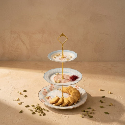 A three-tiered gold metal serving stand with floral-patterned plates, styled with cookies and saffron on a beige textured surface against a matching background.