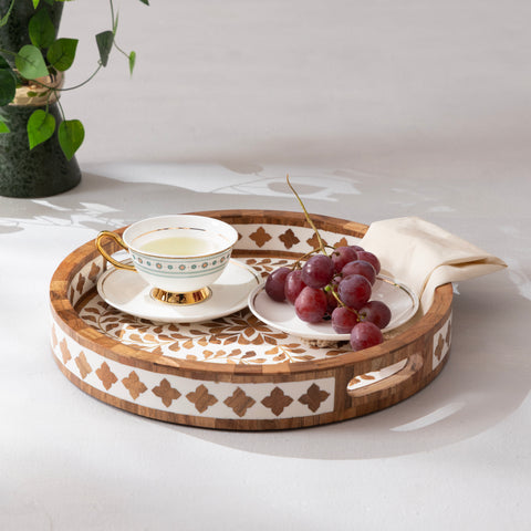 A decorative round wooden serving tray featuring floral patterns with a tea cup, grapes, and a napkin on a light surface.