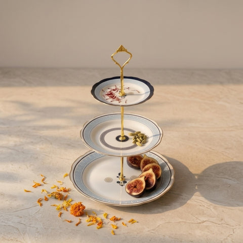 A three-tier gold metal and white ceramic serving stand with blue accents, styled with dried figs and saffron on a beige stone surface under warm sunlight.