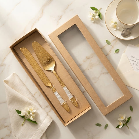 Brass cake knife serving set in a box on a marble surface with flowers and a teacup.