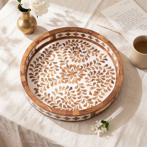Round wooden serving tray with a white inlay featuring a detailed floral and leaf pattern, styled on a beige linen tablecloth with a small vase, handwritten letter, and a cup of tea nearby.