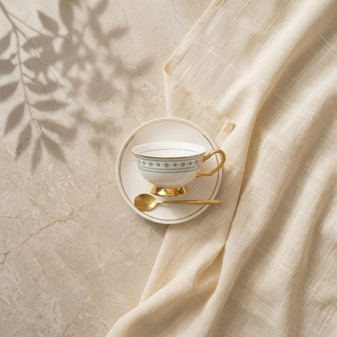Teacup with saucer and spoon on a marble surface with fabric and leaf shadows