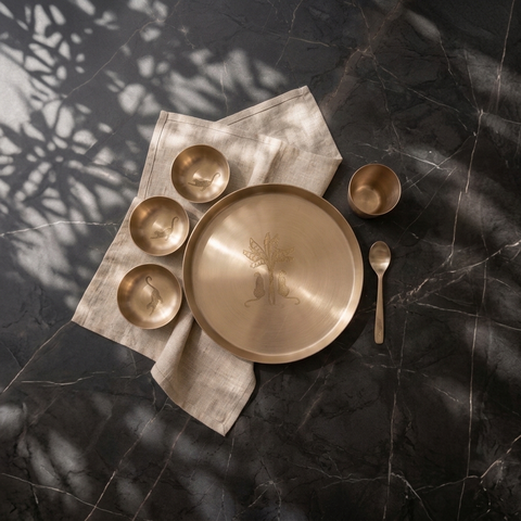 A set of matte brass tableware, including a large platter, four small bowls, a cup, and a spoon, arranged on a beige linen napkin over a dark marble surface with leaf shadows.