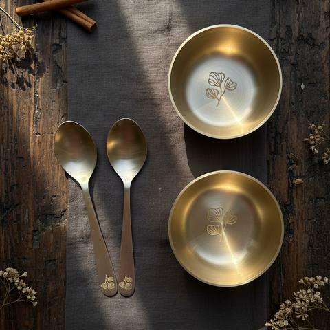 kansa bowl sets and spoons on a wooden surface with cinnamon sticks and dried flowers.