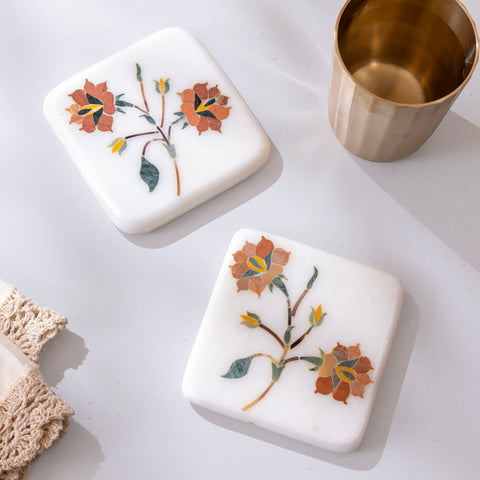 Two square marble coasters with floral designs on a white surface next to a gold cup.