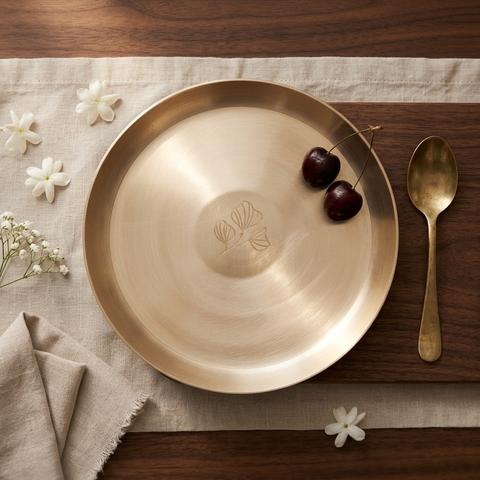 A round brushed brass plate with a brushed metal finish, featuring an engraved lotus flower motif in the center. The plate is styled on a beige linen runner alongside a matching brass spoon, white flowers, and two dark cherries on a dark wood table.