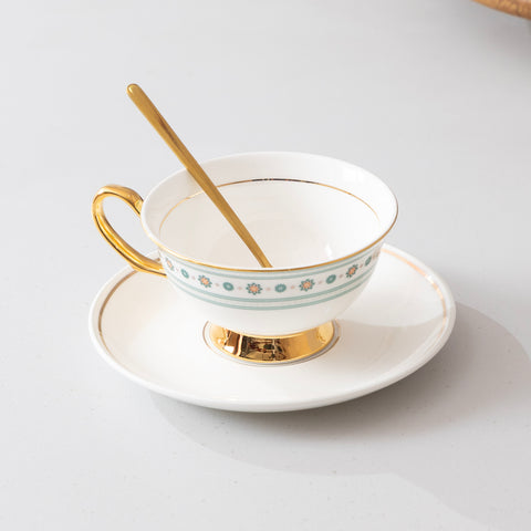 Teacup with floral pattern and gold trim on a matching saucer with a gold spoon on a light gray background