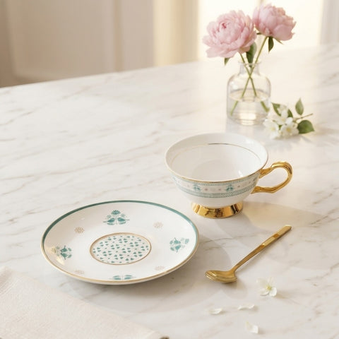 Teacup and saucer set with gold accents on a marble surface, accompanied by a vase of pink flowers.