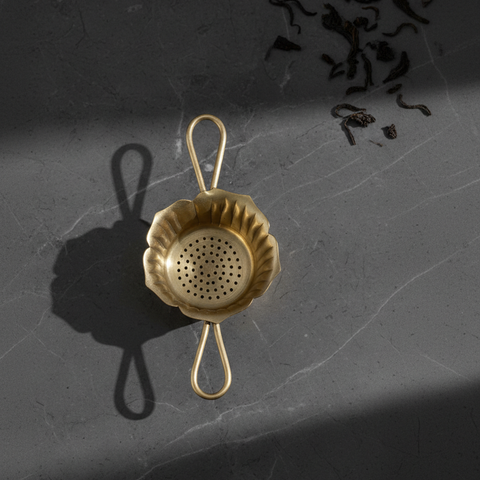 Gold tea strainer on a dark textured surface with tea leaves.