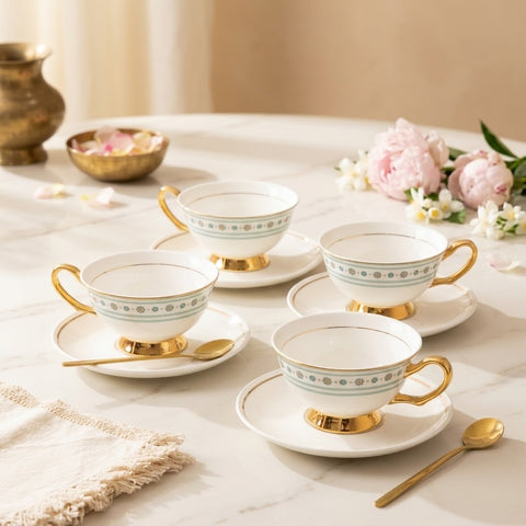 Four white ceramic teacups with gold feet and handles, decorated with a delicate blue and green floral band, arranged on matching saucers on a light marble table. Gold spoons, pink peonies, and a brass vase are visible in the styled setting.