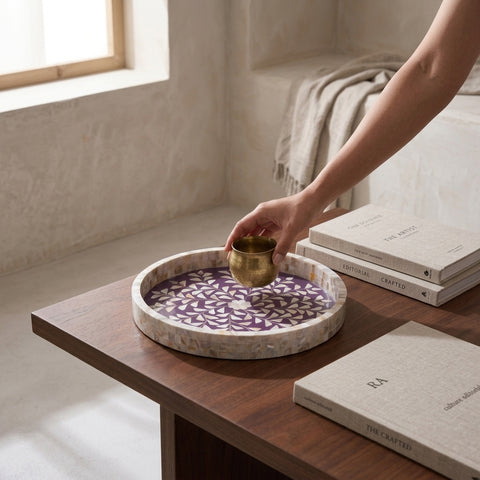 Person holding a brass cup over a violet round tray with mother of pearl inlay on a wooden table with books in the background.